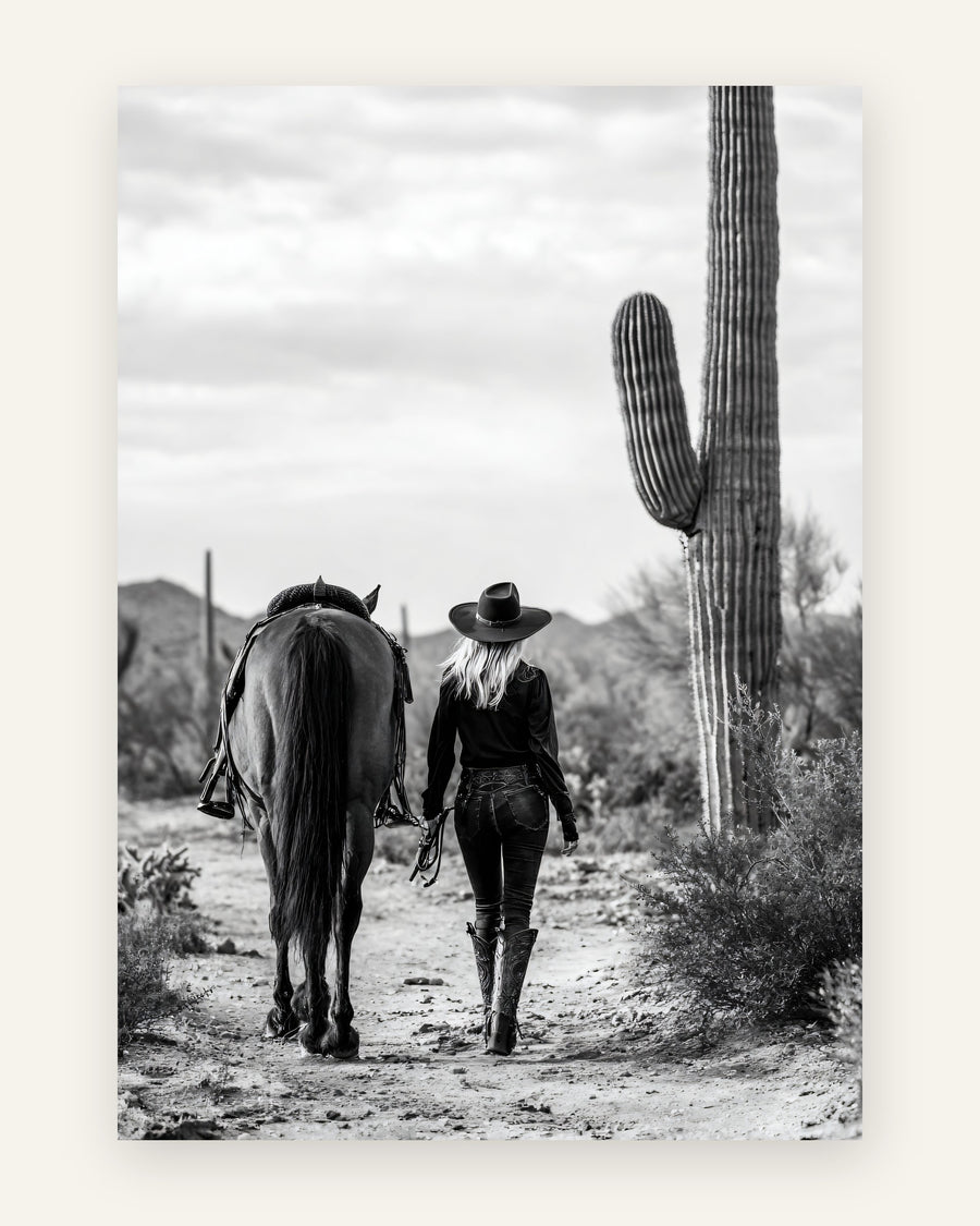 Sort-hvidt fotografi af cowgirl med cowboyhat der går med sin hest ad en ørkensti blandt saguaro-kaktusser