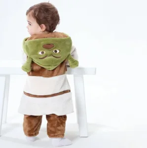 Dette billede viser et nuttet Shrek-kostume til børn, bestående af en blød, lysebrun overdel med vest og matchende grønne bukser. Dragten fuldføres af en karakteristisk grøn hætte med Shreks ikoniske ører, hvilket skaber et sjovt og genkendeligt look til fastelavn eller leg.