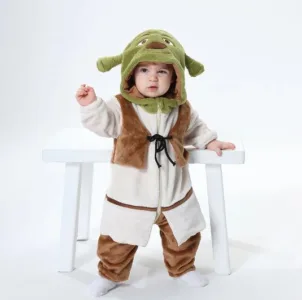 Dette billede viser et sjovt Shrek-kostume til børn, bestående af en beige overdel med påsyet vest, ternede bukser og en grøn maske med de karakteristiske troldeører. Dragten er designet i bløde materialer, der gør den behagelig for småbørn at have på til fastelavn eller leg.