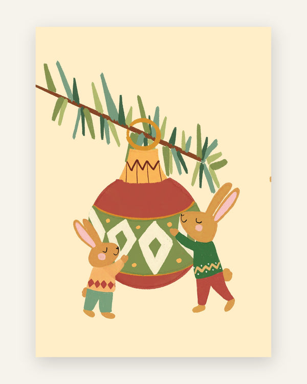 Børneplakat med to brune kaniner i julesweatre, der pynter en stor rød og grøn julekugle på en grangren, på cremefarvet baggrund til børneværelset