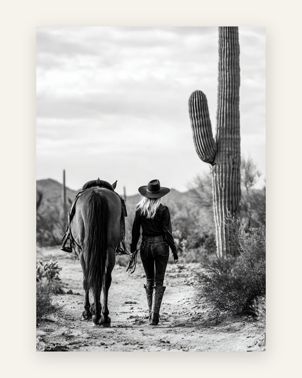 Sort-hvidt fotografi af cowgirl med cowboyhat der går med sin hest ad en ørkensti blandt saguaro-kaktusser